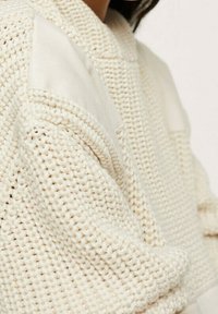 Sweter dziany w kremowym kolorze z teksturowanym wzorem. Posiada gładkie beżowe akcenty na ramionach oraz luźny fason. Okrągły dekolt.