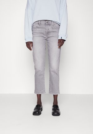 Person trägt hellgraue kurze Jeans, schwarze Leder-Loafers und ein hellblaues langärmliges Oberteil, steht vor einem weißen Hintergrund.