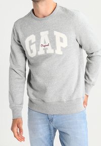 Hombre con sudadera clara de GAP con el texto "Original" debajo del logotipo, combinado con jeans de color azul claro, de pie con una mano en el bolsillo.