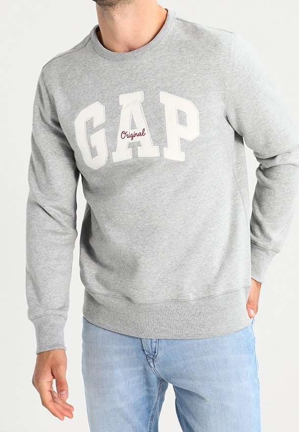 Hombre con sudadera clara de GAP con el texto "Original" debajo del logotipo, combinado con jeans de color azul claro, de pie con una mano en el bolsillo.