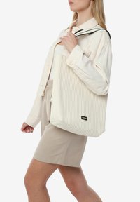 Grande borsa tote bianca realizzata in materiale a righe, con una tracolla nera e un piccolo logo nero sul davanti.