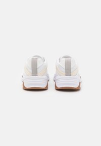 Białe buty sportowe z teksturowaną cholewką, z beżowymi akcentami i brązową gumową podeszwą. Projekt zawiera kontrastujące przeszycia i detal logo.