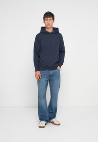 Granatowy hoodie z przednią kieszenią, w połączeniu z jasnoniebieskimi dżinsami w stylu dzwonów i białymi trampkami, stojący na jednolitym białym tle.