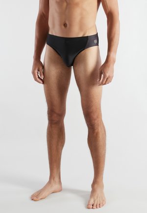 Erwachsener männlicher Model, barfuß stehend, trägt schwarze Badehose mit gemusterten Seitenpanelen und kleinem Logo auf der linken Seite.