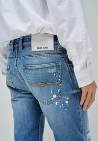 Jeans de denim azul con bolsillos traseros, costuras amarillas y salpicaduras de pintura en blanco y azul. La etiqueta dice "SALSA JEANS."