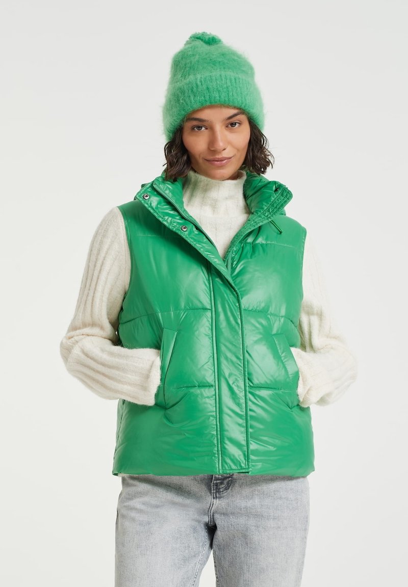 Green glossy puffer vest with a high collar, two large front pockets, and snap closure. Paired with a fluffy green beanie and cream sweater.