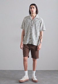 Korte mouwen overhemd met een geometrisch patroon in lichte kleuren, gecombineerd met een bruine short en crèmekleurige sneakers. Casual outfit.