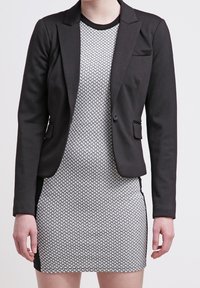 Blazer nero con un design aderente e due tasche frontali, indossato sopra un vestito grigio a fantasia con scollatura tonda e maniche corte.