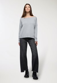 Maglione grigio chiaro a spalle scoperte, dalla vestibilità rilassata, con maniche lunghe. Abbinato a jeans scuri a gamba larga e stivali neri alla caviglia.