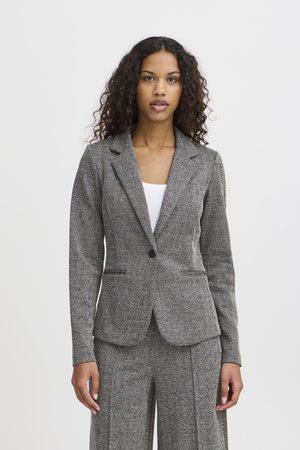 Femme aux cheveux bouclés vêtue d'une veste de costume grise à motif chevrons et d'un pantalon large assorti, par-dessus un haut blanc, debout devant un fond blanc.