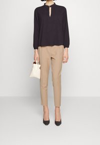 Zwarte blouse met lange mouwen en een keyhole-halslijn met ringdetail, gecombineerd met lichtbeige broek in maatwerk en zwarte pumps met spitse neuzen.