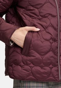 Mano nella tasca di una giacca trapuntata bordeaux con cuciture a forma di cuore, accanto a una cerniera argentata, sopra un paio di pantaloni a quadri.