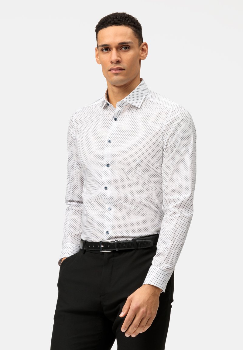 Chemise blanche à longues manches avec un motif de petits pois bleus, col boutonné et boutons bleus, associée à un pantalon noir et une ceinture.