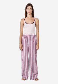 Młoda kobieta stoi barefoot, ubrana w jasnoróżową koszulkę na ramiączkach z ciemnym wykończeniem oraz luźne różowe spodnie z białymi pionowymi paskami.