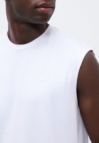 Gros plan d’un homme à la peau foncée portant un débardeur blanc avec un petit logo discret sur la poitrine, regardant sur le côté.