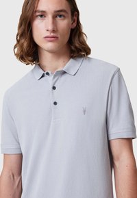 Grå polo skjorte laget av teksturert stoff, med flat krage, tre-knapp placket og en liten brodert logo på brystet.