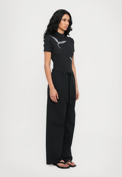 Black fitted top with short sleeves and a high neckline, featuring silver wing patterns. Wide black pants with a tied waistband and flip-flops.