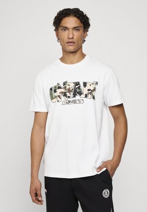 Homme aux cheveux bouclés portant un T-shirt blanc avec texte camouflage "UNFAIR ATHLETICS" et pantalon noir avec logo, debout devant un fond clair uni.