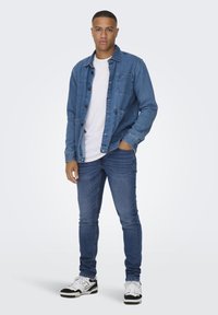 Chaqueta de denim en azul con cierre de botones en la parte delantera y dos bolsillos en el pecho, combinada con jeans ajustados azul y zapatillas en blanco y negro.