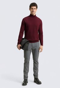 Pull à col roulé bordeaux associé à un pantalon gris sur mesure et des bottines noires. Le mannequin tient un sac noir, debout devant un arrière-plan neutre.