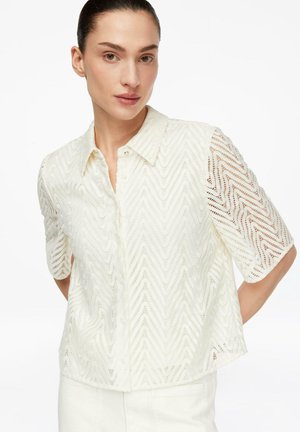 Frau trägt weißes Spitzenhemd mit kurzen Ärmeln und Chevron-Muster, posiert mit den Händen hinter dem Rücken vor neutralem Hintergrund.