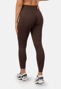 Brązowe legginsy z wysokim stanem, wykonane z elastycznego materiału, o gładkim wykończeniu i dopasowanym kroju, w połączeniu z białymi i szarymi butami sportowymi.