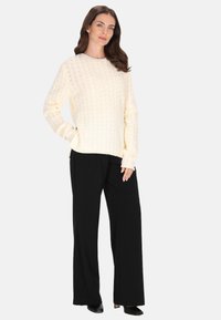 Krém színű, kötött pulóver texturált mintával, kerek nyakkivágással és hosszú ujjakkal, fekete, bő szárú nadrággal és fekete magassarkú cipővel párosítva.
