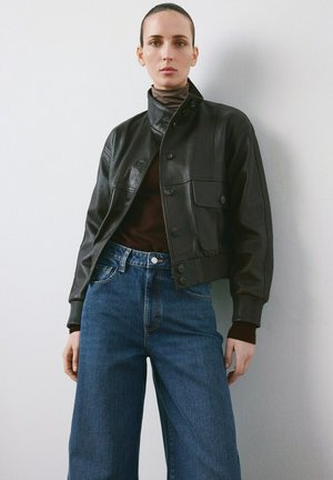 Mujer con chaqueta de cuero negra, suéter de cuello alto marrón y jeans azules de talle alto, de pie contra una pared blanca.