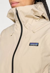 Beez, veekindel jakk kõrge kraega, musta tõmblukuga ja logo plaadi kui sildiga. Omab reguleeritavaid nööre ja siledat tekstuuri.