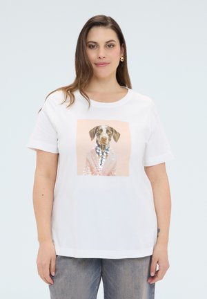 Kvinde iført en hvid t-shirt med en palietbroderet grafik af en hund iført en pink jakke og halskæde, stående mod en ensfarvet baggrund.