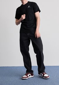T-shirt nera in cotone con logo Jordan bianco, jeans in denim nero con cuciture a contrasto, e sneaker Nike bordeaux e bianche con accenti neri.