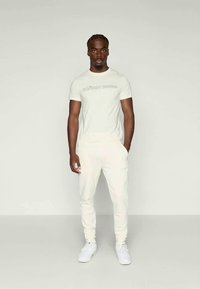 Tricou alb din bumbac cu textul "BUJRN BORG" pe față, asortat cu pantaloni jogger albi și adidași albi. Fundal simplu.
