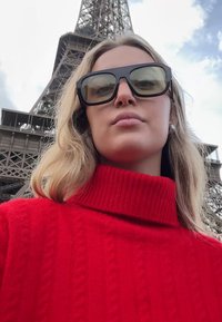 Röd kabelstickad polotröja, oversize-passform, kombinerad med stora, rektangulära svarta solglasögon. Eiffeltornet delvis synligt i bakgrunden.