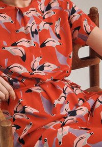 Röd klänning med ett mönster av stiliserade flamingos i nyanser av rosa, lila och svart, med en rynkad midja och korta ärmar.