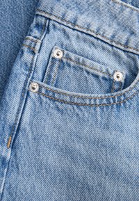 Κοντινή λήψη μπροστινής τσέπης σε ανοιχτό μπλε τζιν με πορτοκαλί ράμματα και ασημένιες τρουκ στις γωνίες της τσέπης.