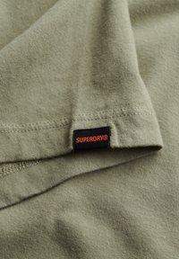 Oliven grøn bomulds T-shirt med et sort mærke med rød "SUPERDRY" tekst. Glat tekstur og korte ærmer med syede kanter.