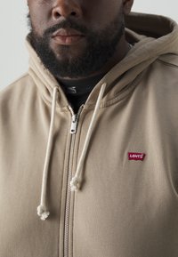 Beige Zip-Hoodie aus weichem Stoff, mit Kapuze und verstellbarem Kordelzug, silbernem Reißverschluss und rotem Levi's Logo auf der linken Brust.