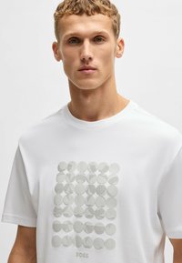 Hvid t-shirt med et geometrisk mønster af cirkulære former i forskellige teksturer. Rund hals og korte ærmer med et diskret logo under designet.
