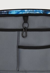 Grå tygväska med svart trim inuti, har flera fickor och en klämma. Övre kanten framhäver ett blått mönstrat foder.