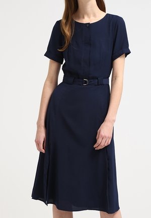 Vrouw draagt een marineblauwe midi-jurk met korte mouwen, een riem, een ronde hals en een knoopsluiting aan de voorkant.