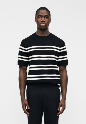 Ung sort mand iført en sort kortærmet skjorte med vandrette hvide striber, stående foran en ensartet hvid baggrund.