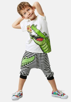 Kindermode mit einem weißen T-Shirt, das ein grünes Krokodil-Graphic zeigt, und gestreiften schwarz-weißen Shorts mit einem Krokodil-Patch. Bunte Sneakers.