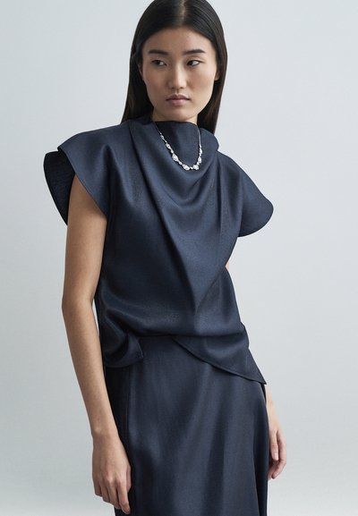 Haut bleu marine avec un col structuré et drapé, manches courtes type casquette, associé à une jupe assortie. Tissu avec une légère brillance ; design simple.