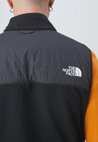 Black and orange vest with a high collar, featuring a smooth upper section and textured lower part. The North Face logo is prominently displayed.