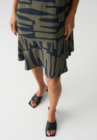 Green and navy patterned dress with a flared hem, textured fabric; paired with black square-toed slide sandals.