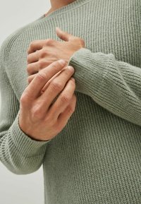 Lichtgroene gebreide trui met textuur, voorzien van geribde manchetten en halslijn. Zichtbare handen die de stof vasthouden, waarmee de zachte textuur en het ontwerp worden getoond.