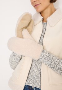 Beige shearling vest med en blød krave, parret med lysebrune vanter. Vanterne har en blød tekstur og en buet form.