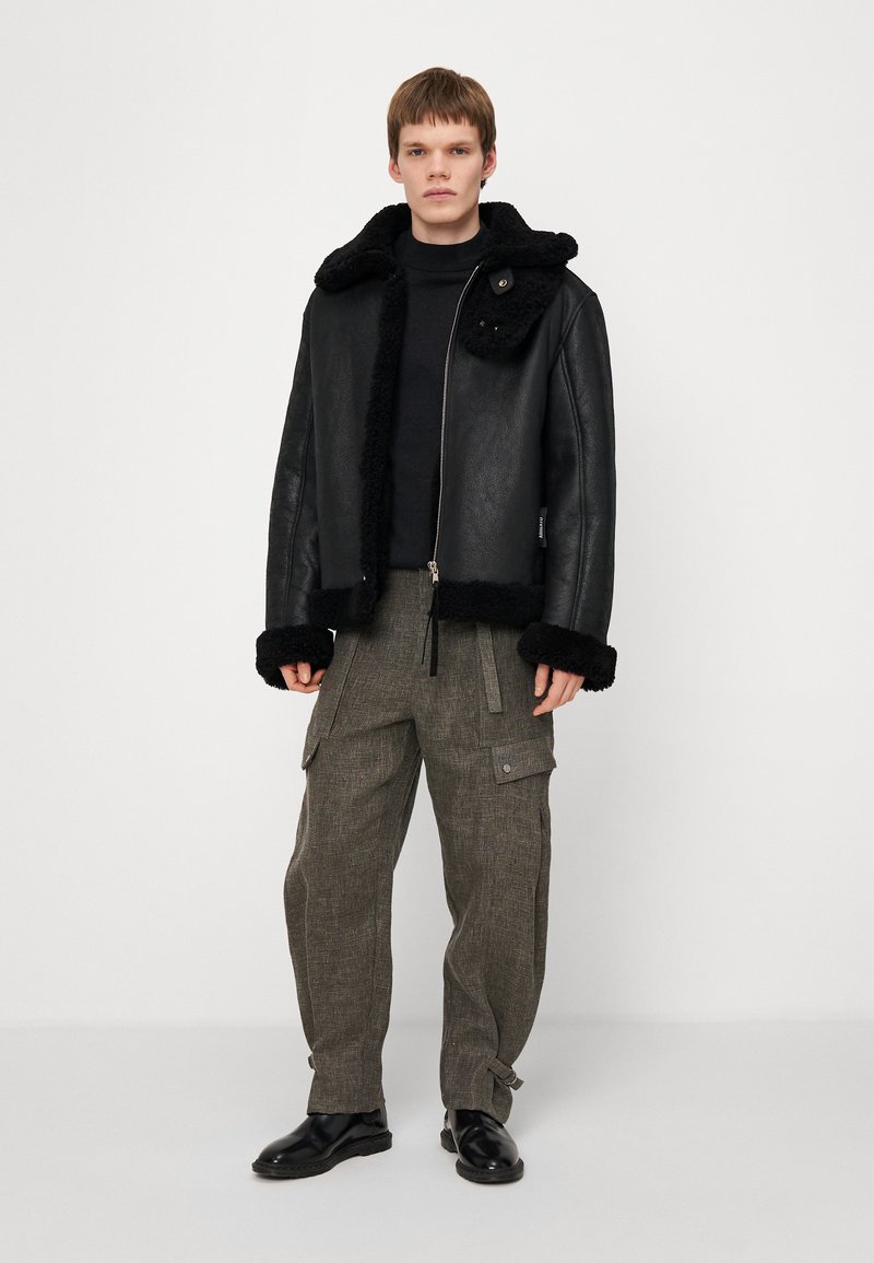 Chaqueta de cuero negra con cuello y puños de piel de oveja, cierre de cremallera, llevada sobre un cuello alto negro y combinada con pantalones cargo grises texturizados.