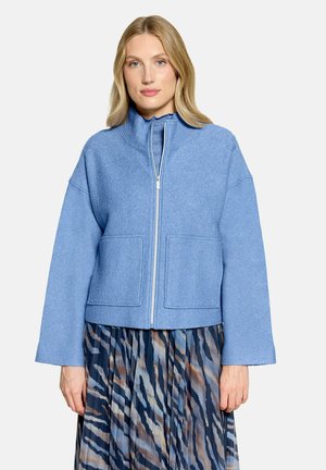 Femme aux cheveux blonds portant une veste zippée bleu clair et une jupe plissée avec des motifs abstraits bleus, noirs et bruns.