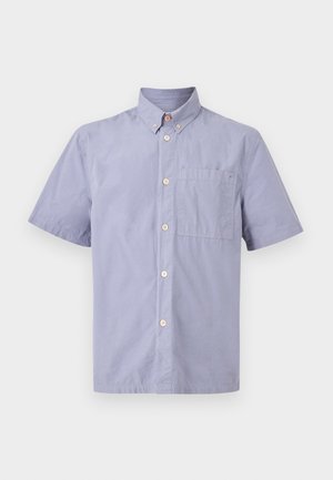 Uomo in piedi che indossa una camicia azzurra a maniche corte con bottoni, pantaloni marroni e scarpe da ginnastica bianche su uno sfondo uniforme.